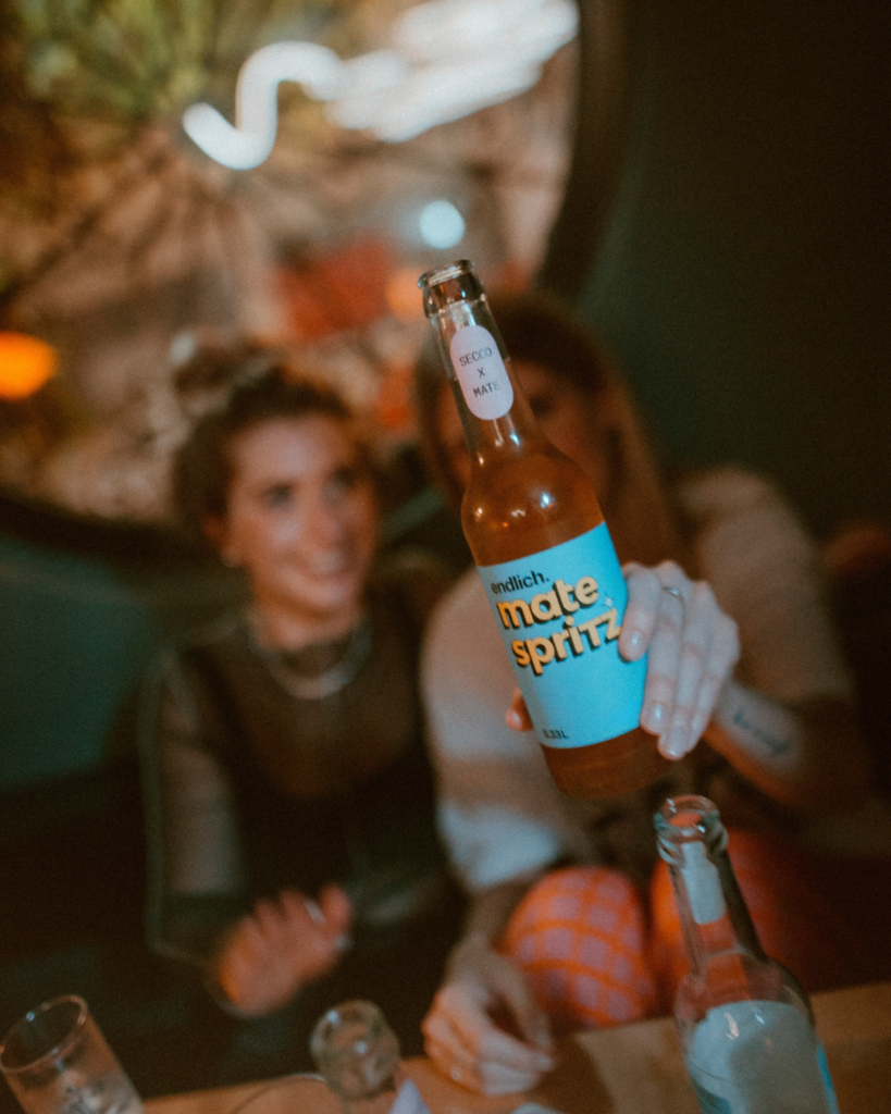 Zwei Frauen sitzen an einem Tisch im Spritz und eine hebt eine Flasche Mate Spritz in die Kamera.